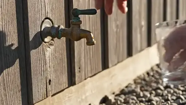A hand filling a clear glass with water at an old brass garden spigot on a shed wall
